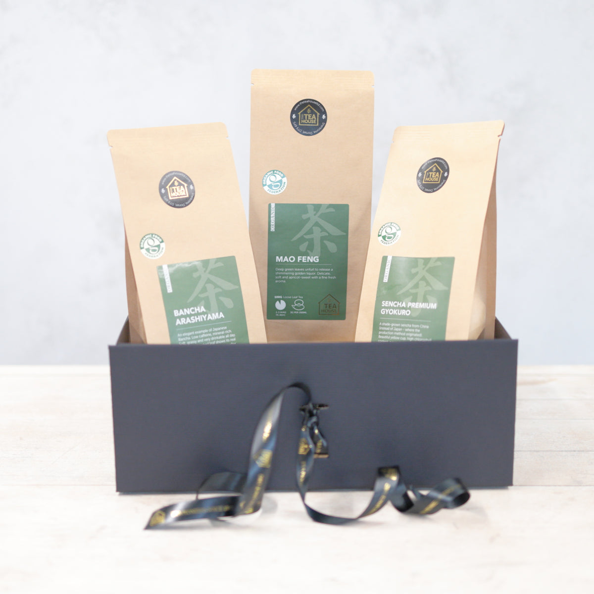 Three 100g packs of organic green tea in a black gift box with black and gold ribbon