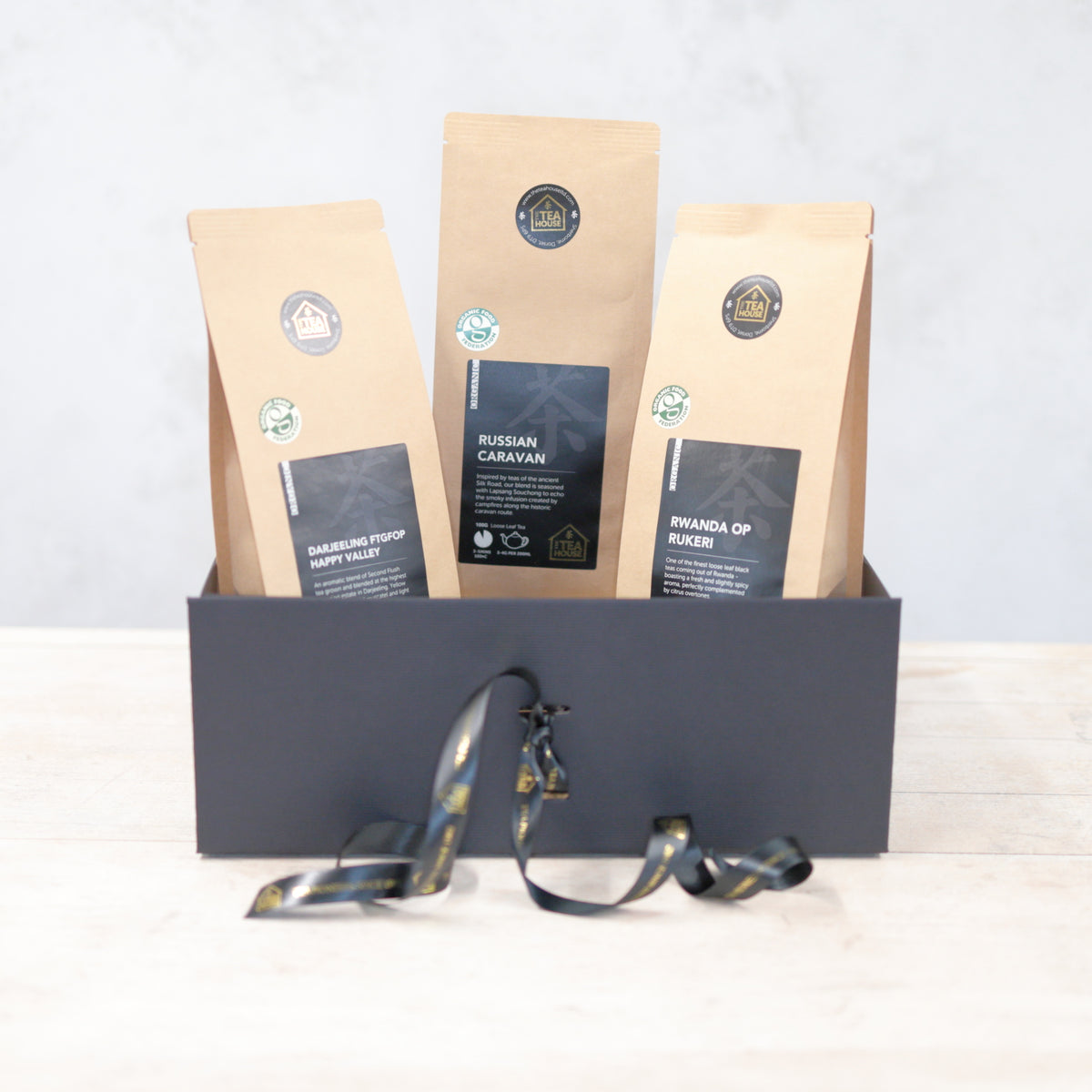 Three 100g packs of organic black tea in a black gift box with black and gold ribbon
