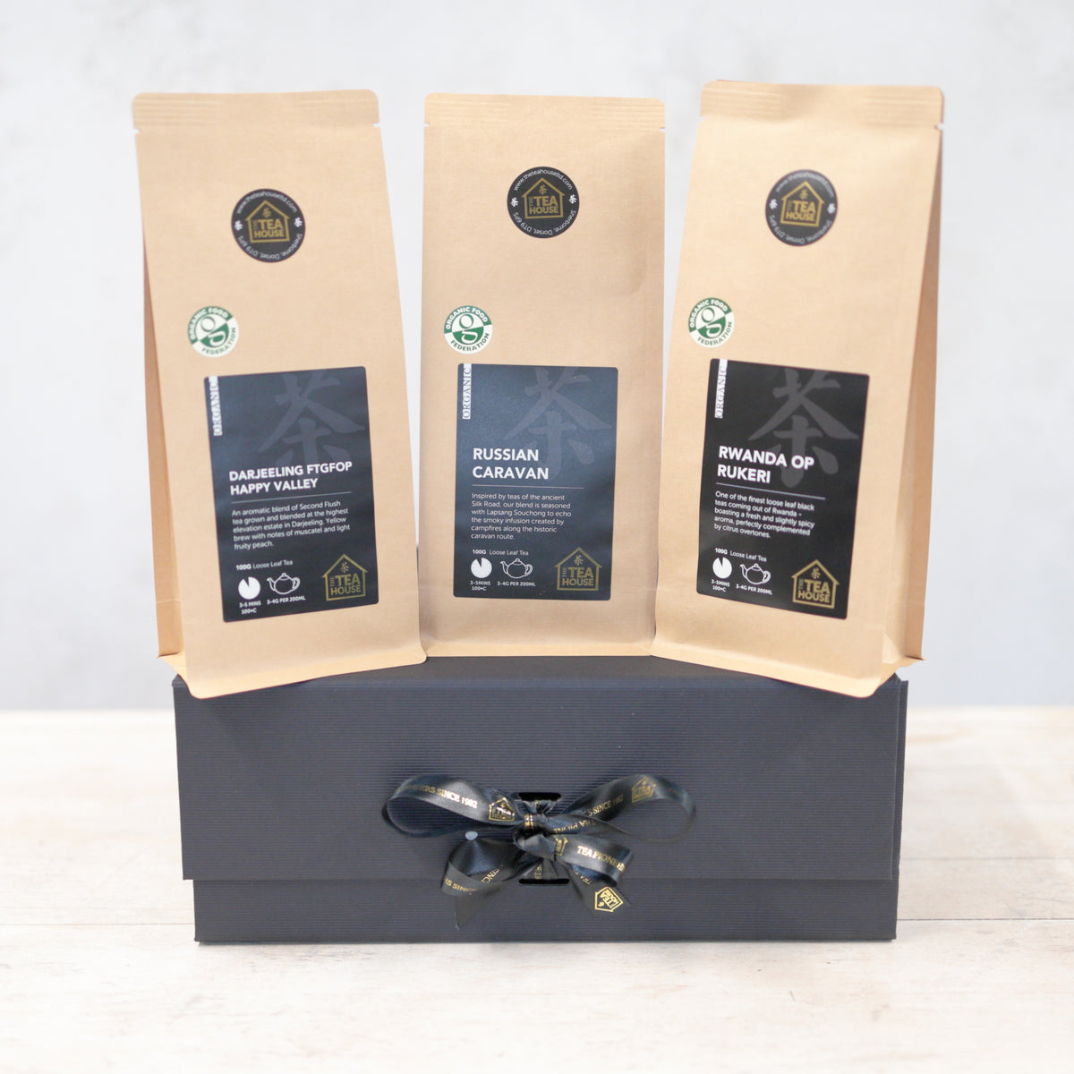 Three 100g packs of organic black tea on a black gift box with black and gold ribbon