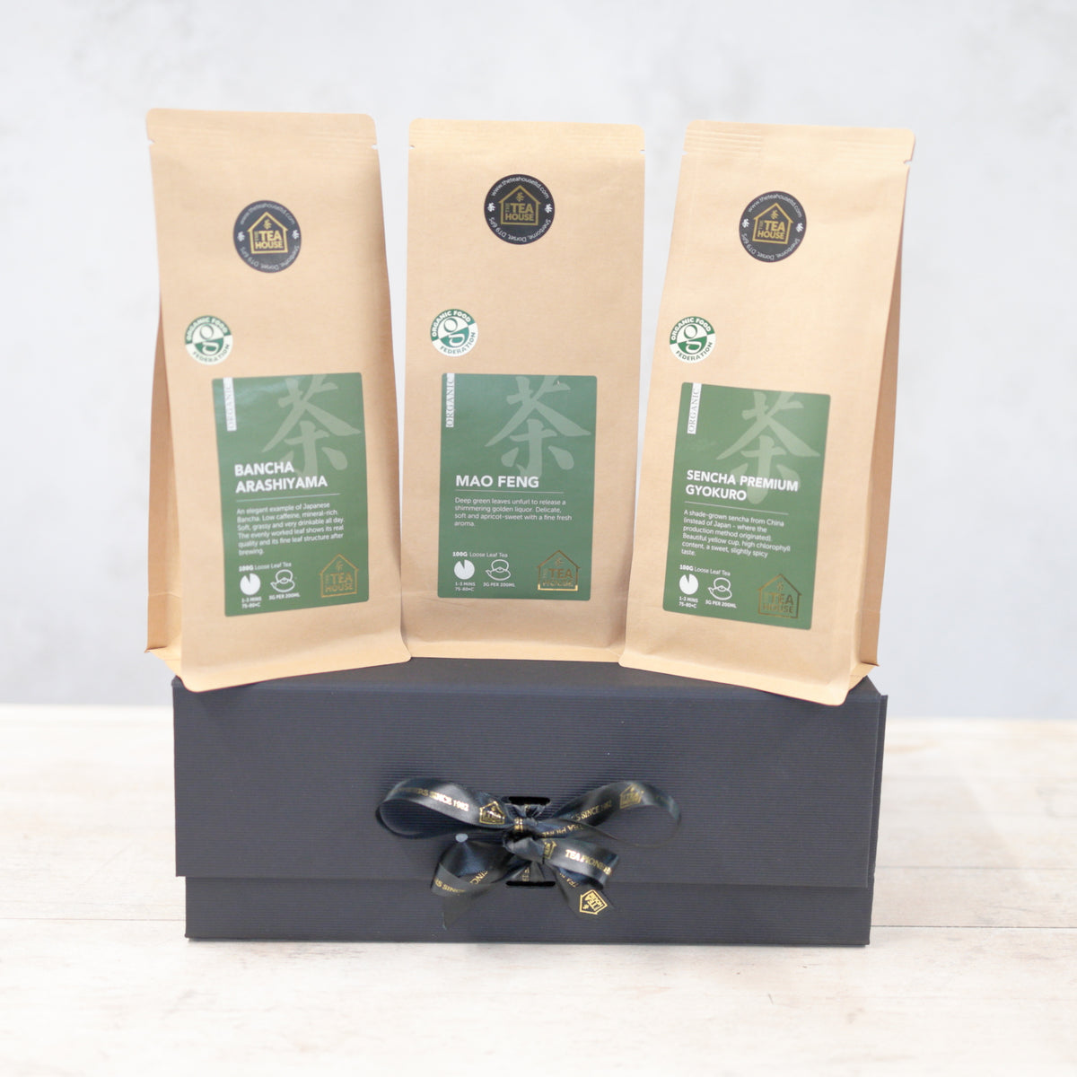 Three 100g packs of organic green tea on a black gift box with black and gold ribbon