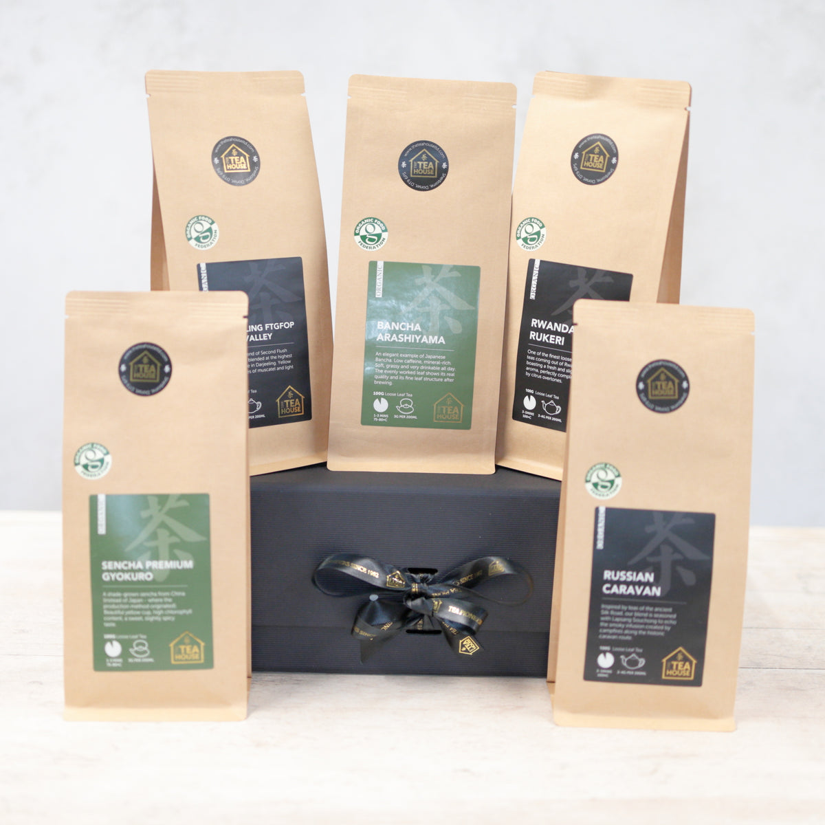 Three 100g packs of organic black tea and two 100g packs of organic green tea on a black gift box with black and gold ribbon  