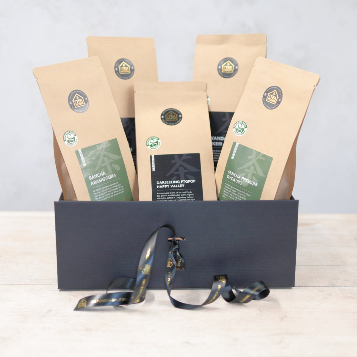 Three 100g packs of organic black tea and two 100g packs of organic green tea in a black gift box with black and gold ribbon